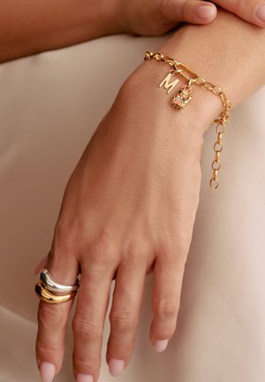 Gold chain bracelet featuring an "M" charm and a decorative charm. Additional silver and gold rings on fingers with a smooth finish.