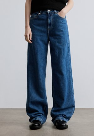 Personne portant un jean bleu ample à jambes larges avec des chaussures noires, une main dans la poche, debout sur un sol gris sur un fond gris clair.