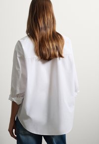Camisa de algodón blanca con mangas arremangadas, corte relajado, dobladillo curvado y detalle en el yugo trasero, combinada con vaqueros azules de lavado medio.