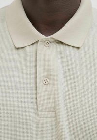 Beige strikket polo-shirt med et tekstureret mønster, der har en blød krave og to beige knapper ved knapstriben.