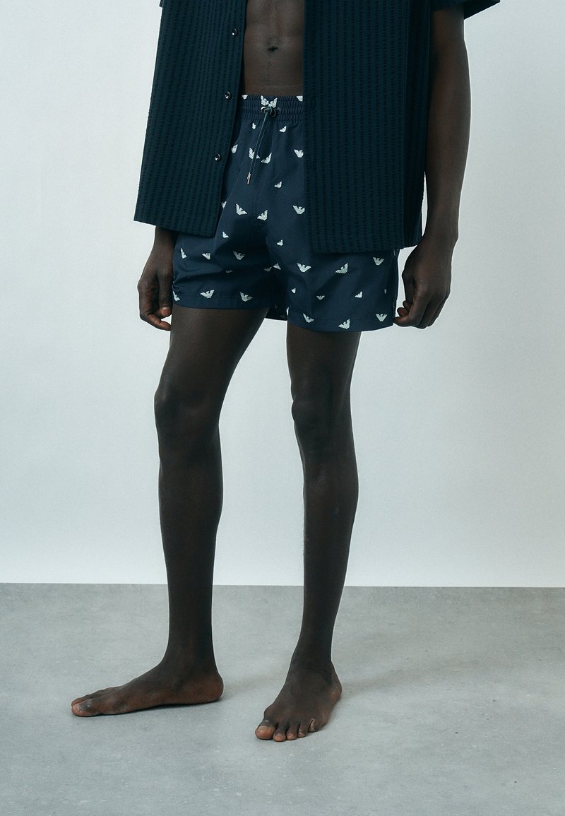 Άνδρας που φοράει ναυτικό σορτς με λευκά λογότυπα αετού και σκουρόχρωμο ανοιχτό πουκάμισο, στέκεται ξυπόλητος σε γκρι δάπεδο μπροστά σε μονόχρωμο τοίχο.