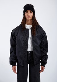 Giacca bomber nera con zip frontale, polsini a coste e tasche laterali. Indossata sopra una camicia bianca e abbinata a pantaloni in denim scuri. Berretto nero.