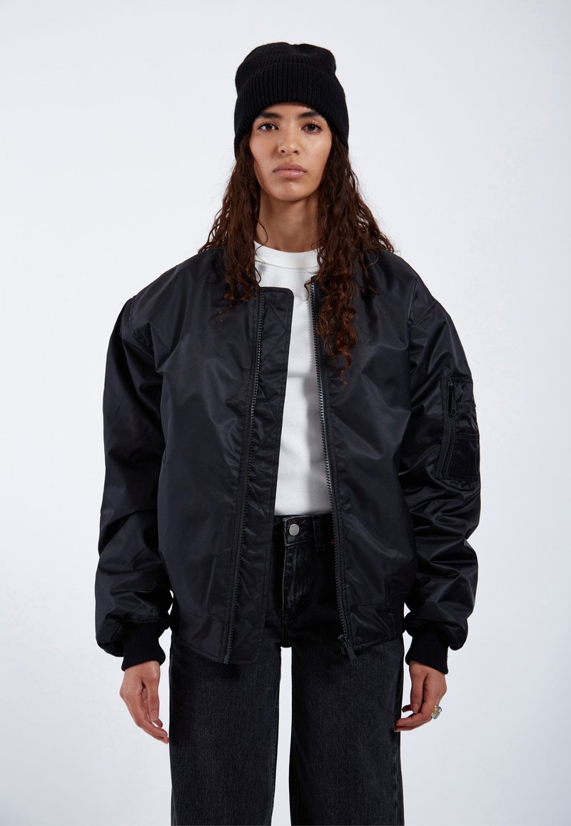 Giacca bomber nera con zip frontale, polsini a coste e tasche laterali. Indossata sopra una camicia bianca e abbinata a pantaloni in denim scuri. Berretto nero.