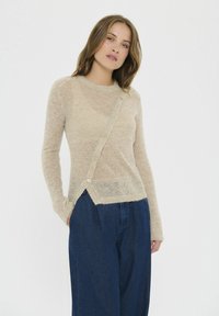 Beige stickad tröja med rundad halsringning, långa ärmar och knappstängning fram. Har en strukturerad yta och en trappad fåll.