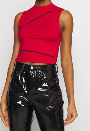 Top court rouge sans manches avec deux fermetures éclair noires diagonales porté avec un pantalon en vinyle noir brillant taille haute.