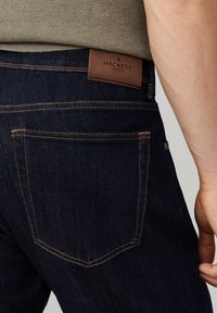 Krupni plan tamnoplavih traperica s narančastim šavovima i smeđom kožnom naljepnicom Hackett London na pojasu, koje nosi osoba u zelenom džemperu.