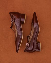 Escarpins en cuir verni marron avec un bout pointu, un petit talon carré et un détail de boucle sur le côté. Texture lisse et finition brillante.