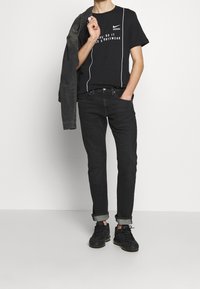 Schwarzes Baumwoll-T-Shirt mit weißem Logo und Text, kombiniert mit dunkelblauen Jeans, hochgerollten Ärmeln und schwarzen Sneakern. Jacke über die Schulter gelegt.