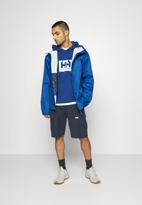 Chaqueta impermeable azul con acentos grises sobre una sudadera con capucha color marino. Combinada con pantalones cortos oscuros y zapatillas deportivas blancas. Detalles del logo sutiles presentes.