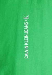 Groene katoenstof met verticale witte tekst: "CALVIN KLEIN JEANS CK," met een gladde textuur en een minimalistisch ontwerp.