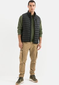 Schwarze Steppweste über einem grünen Pullover, kombiniert mit sandfarbenen Cargo-Hosen und olivgrünen Sneakers. Bemerkenswerte Merkmale sind Reißverschlüsse und Taschen.