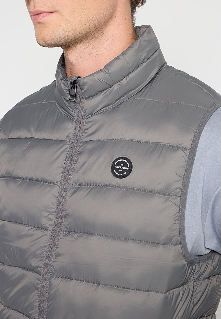 Chaleco acolchado gris con cuello alto, cremallera y costuras horizontales. Presenta un logo circular negro en el pecho. Textura de tela ligera.