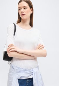 Jeune femme aux cheveux lisses portant un t-shirt blanc, un jean bleu, une chemise à rayures bleu clair nouée autour de la taille et un sac d'épaule noir.