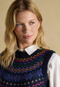 Jūras zilā adīta džemperveste ar krāsainiem ģeometriskiem rakstiem, ar ribu apkakli un pleciem, uzvilkta pār balto krekla ar apkakli.