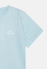 Světle modré bavlněné tričko s kulatým výstřihem, na kterém je vytištěný text "MADS NØRGAARD COPENHAGEN" v bílé barvě poblíž levého prsu.
