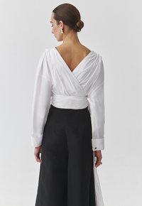 Witte wikkelblouse met een V-achterkant en lange mouwen, gecombineerd met hoge zwarte wijde broek; kenmerken zijn vloeiende zijde stof en manchet details.