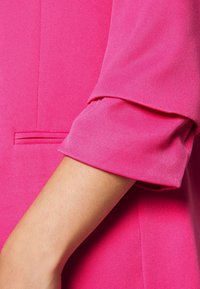 Giacca su misura fucsia con maniche a cuffia ripiegata e un piccolo dettaglio di tasca orizzontale sul braccio. Tessuto dalla superficie liscia.