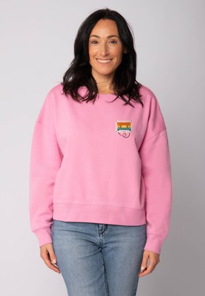 Glückliche Frau mit dunklem Haar trägt einen rosa Sweatshirt mit einem kleinen, bunten Kassettenton-Design, kombiniert mit hellblauen Jeans.