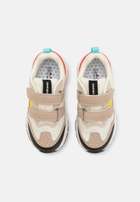 Adidași pentru copii bej și albi cu două bare velcro, accente negre și roșii, și tălpi albastre, văzuți din sus pe un fundal alb.