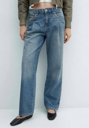 Personne portant un jean bleu délavé à jambes droites, une veste olive verte courte, et des chaussures plates noires à bout carré, debout devant un fond blanc.