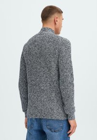 Pulover, teksturiran pletenin v odtenkih sive, z visokim ovratnikom, gubanimi manšetami in robom, sproščen kroj, priložnostno kombiniran z modrimi jeans hlačami.