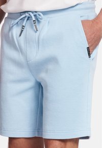 Light blue shorts made from textured material. Features an elastic waistband with drawstrings and side pockets. Branding tag on the side.