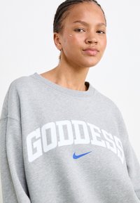 Grijze sweatshirt met het woord "GODDESS" in het wit en een blauwe Nike swoosh. De stof lijkt zacht te zijn met een ronde hals en een relaxed fit.