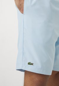 Svetlo modri šorts iz prepustnega materiala, ki se ponaša z diskretno teksturo in vezenim zelenim logojem krokodila na robu.