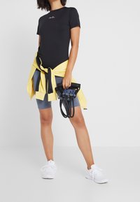 Svart sportt-shirt med logotyp, gul hoodie knuten i midjan, grå cykelshorts, vita sneakers och ett svart motståndsband i handen.