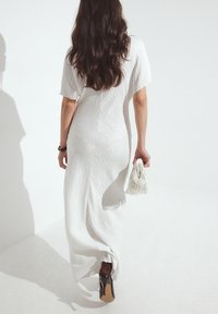 Bela, strukturirana maksi obleka s kratkimi rokavi, ki ima oprijet pas in razpoložen silhueto. Model drži majhno belo torbico s frfotavimi robovi.