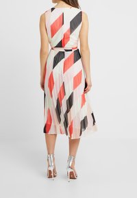 Mujer vestida con un vestido midi sin mangas, plisado, con rayas diagonales en coral, negro y blanco, combinado con sandalias de tacón alto plateadas.