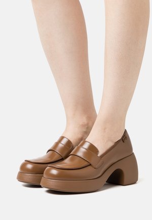 Mocassins à plateforme en cuir marron avec talons épais portés pieds nus sur un fond blanc uni.