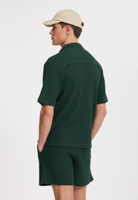 Set de cămașă scurtă cu mâneci scurte și pantaloni scurți texturat, de culoare verde, cu guler. Asortat cu o șapcă de un bej deschis. Ambele articole au o croială lejeră.