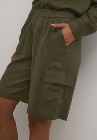Shorts taille haute vert olive avec taille élastique et poche cargo latérale, portés avec une main dans la poche avant droite.