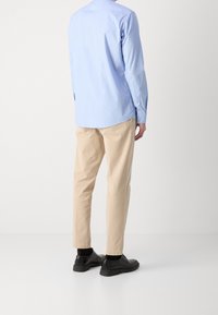 Camicia azzurra chiara con bottoni e maniche lunghe, abbinata a pantaloni chinos beige e scarpe nere, vista dal retro. Design semplice e classico.