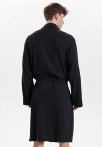 Batín negro de punto waffle con mangas largas, cintura ceñida por cinturón y un dobladillo de longitud media. Presenta una tela texturizada y un diseño de cuello clásico.