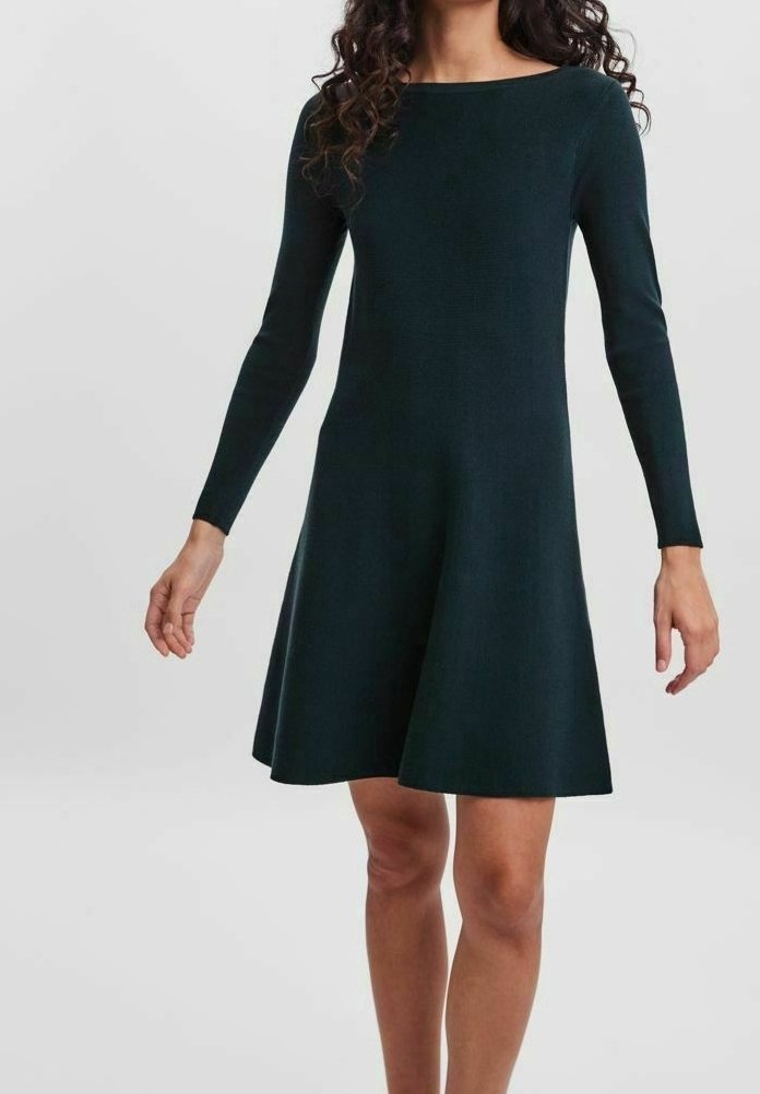 Femme portant une robe A-line à manches longues, de couleur vert foncé, arrivant au genou, avec un décolleté bateau, sur un fond uni.