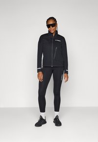 Veste noire courte avec zip, motif en relief et logo. Leggings noirs ajustés, chaussures noires et chaussettes blanches. Des lunettes de soleil complètent le look.