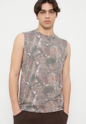 Mladić u bezrukavnoj majici s uzorkom u kamuflaži i tamnim hlačama, stoji s rukama u džepovima na neutralnoj pozadini.