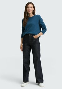 Blauer Strickpullover, hoch taillierte dunkle Jeans mit orangefarbenen Nähten und weiße Sneakers. Modell steht mit einer Hand in der Tasche.