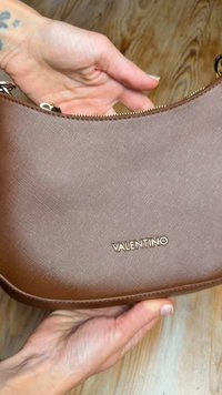 Καφέ τσάντα χειρός Valentino με υφή υλικού, χρυσά εξαρτήματα και λεπτομέρεια λογοτύπου. Το σχήμα είναι στρογγυλεμένο με επάνω κλείσιμο με φερμουάρ.