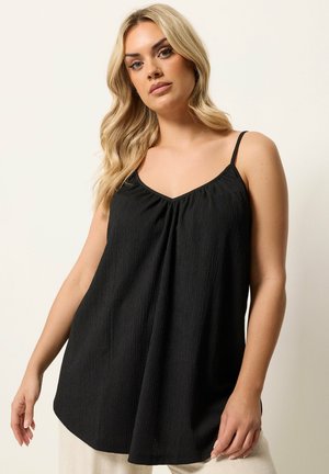 Blond kvinde med løse bølger iført en ærmeløs sort tekstureret top med spaghetti stropper mod en neutral baggrund.