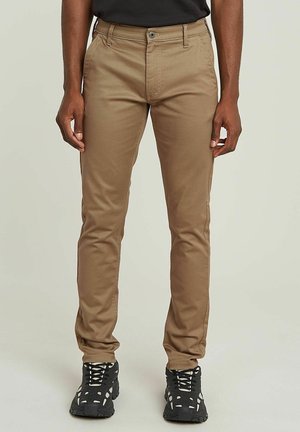 Beige chinos with a slim fit, made from a smooth fabric, featuring a front button closure, belt loops, and side pockets. Worn with black sneakers.