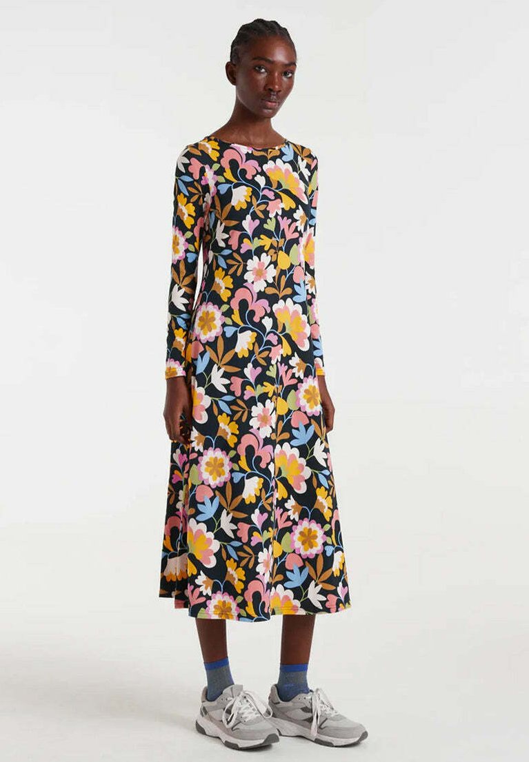 Mujer vistiendo un vestido midi negro de manga larga con un patrón floral colorido, combinado con calcetines azules y zapatillas blancas, de pie contra un fondo liso.