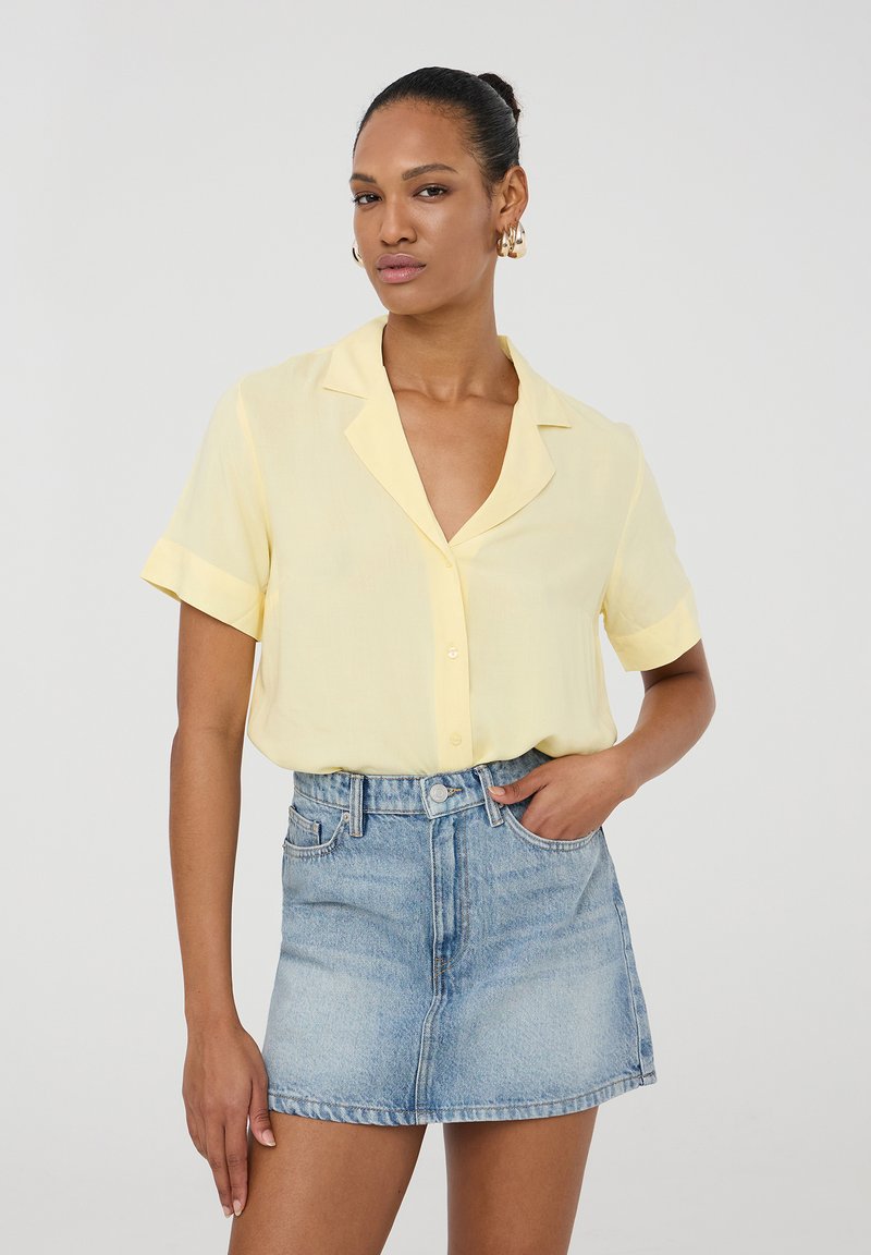 Camicia gialla chiaro a maniche corte con colletto, abbinata a una mini gonna in denim scolorito. Tessuto morbido, vestibilità rilassata, design semplice.