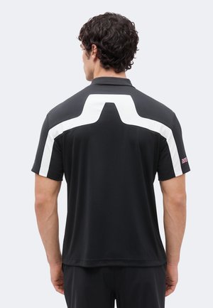 Hombre con camisa negra de manga corta con una distintiva franja blanca angular que atraviesa los hombros y la parte superior de la espalda, visto desde atrás.