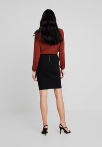 Gonna nera a matita con una texture liscia e un dettaglio con zip argento sul retro, abbinata a una blusa a maniche lunghe di colore ruggine.