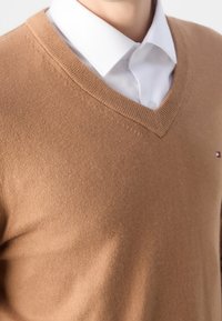 Homme portant un pull à col en V couleur chameau par-dessus une chemise blanche à col, avec un petit logo sur la poitrine.