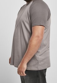 Profilbild av en man i grå kortärmad t-shirt och mörka jeans mot en enfärgad bakgrund.