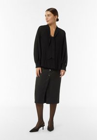 Schwarze plissierte Bluse mit einem Bindekragen, kombiniert mit einem knielangen schwarzen Rock mit Knopfakzenten und hohen Schuhen.